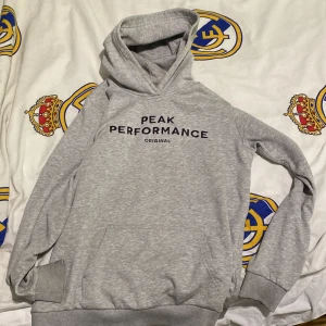 Grå hoodie från Peak Performance - Säljer en stilren grå hoodie från Peak Performance med svart tryck på bröstet. Tröjan har huva, magficka och ribbade muddar vid ärmslut och nederkant. Perfekt för dig som gillar en enkel och sportig look. Nypris: 800. Hör av er vid funderingar!