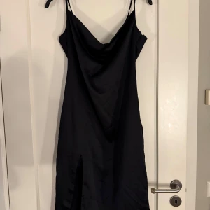 Svart satinklänning med slit - Svart midiklänning i satin med snygg draperad urringning från NA-KD x Nicci Hernestig. Klänningen har en slits framtill och är tillverkad i ett glansigt, satinliknande material som ger en elegant look. Endast använd en gång!