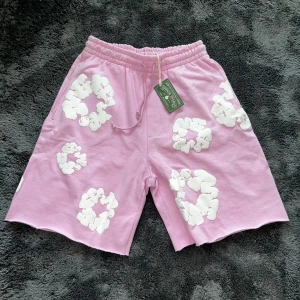 Denim tears shorts. - Säljer ett par rosa shorts från Denim Tears med vita blommiga tryck över hela plagget. De har elastisk midja med snörning och är tillverkade i mjuk bomull. Perfekta för en chill och trendig sommarstil.