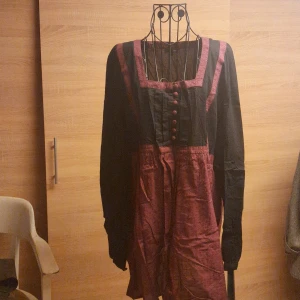 Vinröd och svart klänning By Elois - Unik kortklänning från By Elois i vinrött och svart med långärmad topp och markerad midja. Klänningen har dekorativa knappar framtill, fyrkantig ringning och kontrasterande detaljer. Perfekt för dig som vill sticka ut med en cool och annorlunda look.