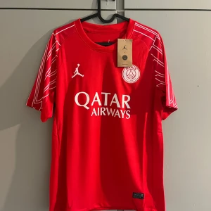 Röd PSG fotbollströja Jordan x Nike - Snygg röd PSG fotbollströja med vita detaljer och tryck från Qatar Airways. Tröjan har korta ärmar med vita grafiska linjer på axlarna och klassisk rund hals. PSG-logga och Jumpman-logga på bröstet. Tillverkad i lätt och ventilerande material.
