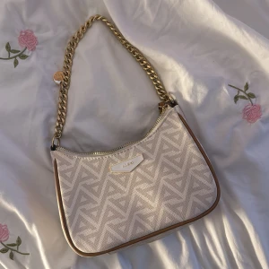 Beige axelväska från ALDO med kedja - Snygg beige axelväska från ALDO med mönstrad canvas och stilrena bruna kanter. Väskan har ett geometriskt mönster, guldfärgad kedjerem och dragkedja upptill. Perfekt storlek för det viktigaste och ger en trendig touch till din outfit.