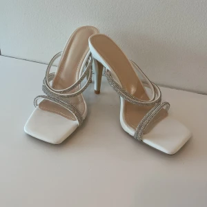 Vita klackade sandaler med strass - Snygga vita sandaler med hög klack och fyrkantig tå. Remmarna är täckta av glittrande strass som ger en lyxig känsla. Perfekta för dig som vill sticka ut med eleganta detaljer och stilren design. Helt nya aldrig använda bara provade köpta för 400kr