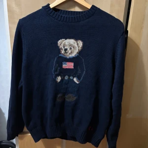 Mörkblå stickad tröja Polo Bear - Mörkblå stickad tröja från Polo Ralph Lauren med ikoniska Polo Bear broderad framtill. Tröjan har rund hals, långa ärmar och RL-broderi nertill. Perfekt för dig som gillar klassisk preppy stil med en twist.
