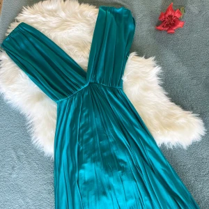 Turkos långklänning med v-ringning - Snygg turkos långklänning i glansigt material med djup v-ringning och breda axelband. Klänningen har ett flytande fall och markerad midja, perfekt för dig som vill sticka ut. Tyget är mjukt och har en lyxig känsla.