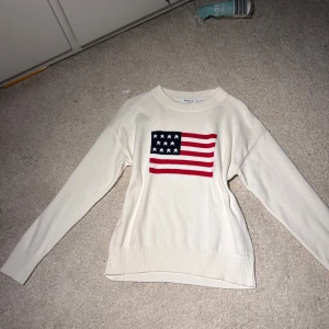 Vit stickad tröja med USA-flagga RYVLS - Vit stickad tröja från RYVLS med lång ärm och ribbade muddar. På bröstet finns en stor USA-flagga i blått, rött och vitt. Tröjan har en klassisk rund halsringning och är perfekt för dig som gillar preppy stil.