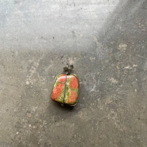 Halsbandshänge med grön och röd sten - Unikt halsbandshänge med en rektangulär sten i grönt och rött, infattad i en silverfärgad metallram. Stenen har ett naturligt mönster och infattningen har en cool korsad detalj på baksidan. Perfekt för dig som gillar färg och naturinspirerade smycken.