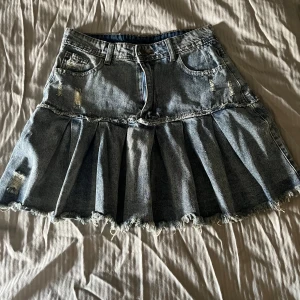 Plisserad jeanskjol med slitna detaljer - Fin kort jeanskjol med plisserad nederdel och råa, fransiga kanter. Kjolen har slitna detaljer och klassiska fickor fram och bak. Perfekt för dig som vill ha en edgy och trendig look. Pris går diskuteras