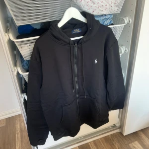 Svart hoodie från Polo Ralph Lauren - Svart hoodie från Polo Ralph Lauren med dragkedja, huva och snörning. Klassisk vit logga broderad på bröstet och stora fickor framtill. Skön passform och perfekt för chill dagar. Materialet är mjuk bomullsmix som känns riktigt nice mot huden. a kopia 