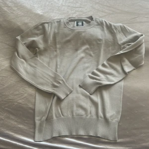 Beige stickad tröja från G-Star RAW - Snygg beige stickad tröja från G-Star RAW med diskret broderad logga på bröstet. Tröjan har rund halsringning, långa ärmar och breda ribbade muddar vid ärmslut och nederkant. Perfekt för lager-på-lager och stilren look.