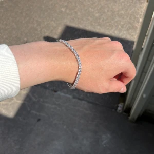 Glittrigt silver tennisarmband - Snyggt tennisarmband i silverfärg med gnistrande stenar runt hela bandet. Armbandet har en klassisk, tunn design och stängs med ett smidigt lås. Perfekt för dig som gillar stilrena och glittriga accessoarer som verkligen syns.