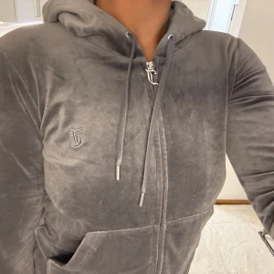 Grå velourjacka från Juicy Couture - Mjuk och snygg grå velourjacka från Juicy Couture med huva, dragkedja och snörning. Klassisk JC-logga broderad på bröstet och stora fickor framtill.