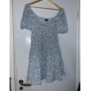 Blommig blå kortklänning Gina Tricot - Supersöt blå kortklänning från Gina Tricot med vita blommor, puffärmar och v-ringning. Klänningen har en luftig passform och är perfekt för varma dagar. Den har en lätt volang nertill och är tillverkad i ett mjukt, tunt material. Aldrig använd, alltså bra skick!😊
