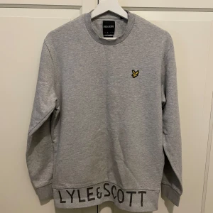 Lyle & Scott sweatshirt  - Snygg grå sweatshirt från Lyle & Scott med klassisk rund hals och ribbade muddar. Tröjan har det ikoniska gula fågel-loggan på bröstet och stor svart logotyp längs nederkanten. Perfekt för en chill och stilren look.