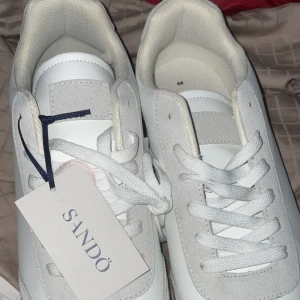 Vita sneakers från Sandö med mockadetaljer - HELT NYA!Snygga vita sneakers från Sandö med ovandel i slätt skinn och ljusa mockadetaljer. Skorna har klassisk snörning och en brun gummisula som ger en stilren kontrast. Perfekta för dig som gillar en clean och fräsch look.