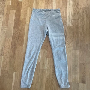 Grå/beige leggings från Stronger, strl S - Snygga ljusgrå/beige leggings från Stronger med hög midja och två vita ränder på ena benet. Tillverkade i stretchigt material med 87% polyester och 13% elastan, perfekta för träning eller chill. Diskret logga framtill och bekväm passform.