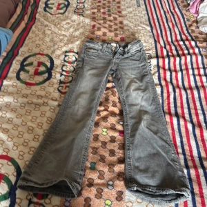 Grå bootcut jeans från Gina Tricot - Snygga grå jeans från Gina Tricot i bootcut-modell. Jeansen har dekorativa bakfickor med broderade detaljer och silvriga knappar. Klassisk femficksdesign och tillverkade i mjukt denimtyg. Perfekta för dig som gillar en avslappnad men trendig look.