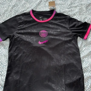 Svart PSG fotbollströja Nike - Snygg svart PSG fotbollströja från Nike med diskret blommönster i tyget. Tröjan har rosa och lila detaljer vid hals och ärmslut samt PSG-logga och Nike swoosh i rosa framtill. Tillverkad i lätt och ventilerande material, perfekt för träning eller match.
