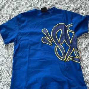 Blå t-shirt från Syna world - Blå t-shirt från Syna world med stor grafisk print i mörkblått och gult som täcker både fram- och baksidan. Klassisk rund hals och korta ärmar. Skön bomullskvalitet och loose fit för en chill vibe. Alla accessoarer finns