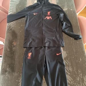 Nike Liverpool  - Säljer en svart Nike Liverpool. Den har hel dragkedja, huva och röda loggor från Nike, Liverpool FC och AXA på bröstet. Tillverkad i lätt och smidigt syntetmaterial, perfekt för fotbollsträning eller chill.