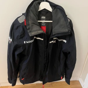 Svart vindjacka från Helly Hansen - Svart vindjacka från Helly Hansen med grå fleecefoder i kragen och vita detaljer samt logga på bröstet och ärmen. Jackan har dragkedja, hög krage och röda detaljer på dragkedjorna. Har lite defekter se bilder 