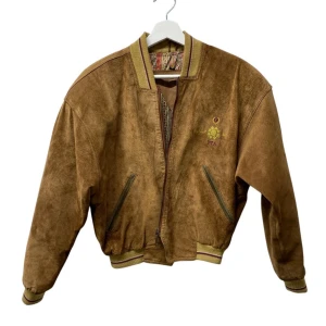 Vintage brun skinnjacka från PTA, storlek S - Snygg brun bomberjacka i äkta skinn från PTA med broderad text på ryggen och coolt paisleymönstrat innerfoder. Jackan har dragkedja, ribbade muddar och två fickor framtill. Med en unik vintagekänsla.