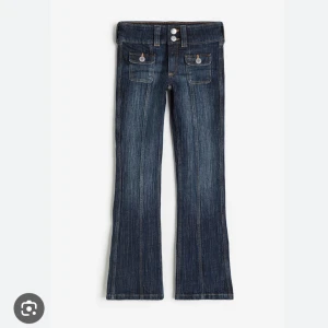 Bootcut jeans mörk denimblå - Mörk denimblå bootcut jeans med snygga pressveck och dubbla knappar i midjan. Framtill finns två fickor med lock och knappdetaljer. Jeansen har breda hällor och klassisk söm längs benen. Perfekta för en cool och avslappnad look.
