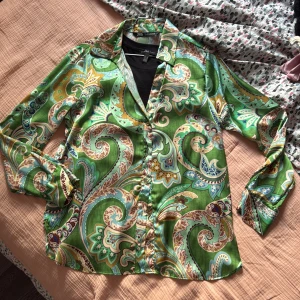 Grön paisleymönstrad skjorta från Zara - Färgstark skjorta från Zara i storlek S med grönt paisleymönster i blått, beige och brunt. Skjortan har klassisk krage, knäppning framtill och långa ärmar. Materialet känns lätt och svalt, perfekt för dig som gillar statement-plagg.