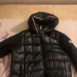 Svart dunjacka från Moncler med huva - Snygg svart dunjacka från Moncler med quiltad front och huva med rödvit detalj. Ärmarna är ribbstickade och jackan har dragkedja framtill. Perfekt för kyliga dagar och ger en sportig men stilren look.