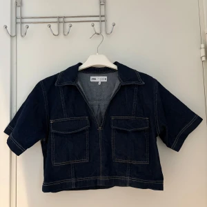 Mörkblå croppad jeansjacka från Zara - Säljer en mörkblå croppad jeansshirt från Zara med kort ärm, två stora bröstfickor och markerade sömmar.