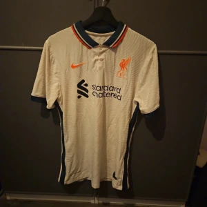 Liverpool FC bortatröja Nike 2021/22 - Liverpool FC:s officiella bortatröja från säsongen 2021/22. Tröjan är vit med mörkblå och orange detaljer på kragen, korta ärmar och broderad Nike-logga. Tillverkad i lätt och ventilerande funktionsmaterial, perfekt för match eller träning.