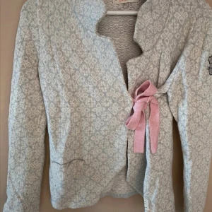 Mönstrad cardigan jacka med rosett - Superfin ljusgrå cardigan jacka med diskret mönster och en söt rosa rosett som knytning framtill. Jackan har lång ärm och en mjuk, stickad känsla – perfekt för lager på lager och chillare dagar.