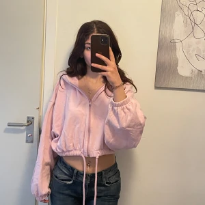 Ljusrosa croppad hoodiejacka - Säljer en ljusrosa croppad hoodiejacka med dragkedja framtill och snörning i midjan. Jackan har oversized passform, långa ärmar med resår vid handlederna och en stor huva. Perfekt för en chill och avslappnad stil.