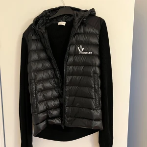 Svart jacka från Moncler - Snygg svart jacka från Moncler med huva och vit logga på bröstet. Jackan är quiltad och har dragkedja framtill samt två fickor med dragkedja. Perfekt att slänga över en hoodie eller tröja för en cool look.