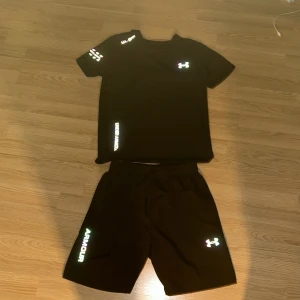 Svart set från Under Armour - Finns även i navy blue och grå. Svart set från Under Armour med elastisk midja och reflekterande logga. Lätta och luftiga i syntetmaterial, perfekta för intensiva träningspass. Snyggt tryck med ARMOUR längs benet ger sportig vibe.