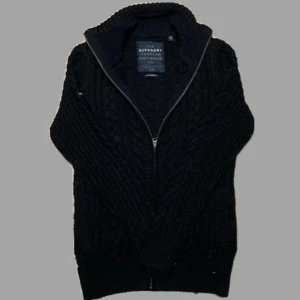 Svart stickad kofta från Superdry - Säljer en svart stickad kofta från Superdry med dragkedja framtill. Koftan har ett snyggt flätmönster och hög krage, perfekt för kyliga dagar. Den är långärmad och har ribbade muddar vid ärmslut och nederkant.