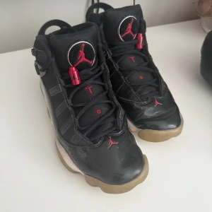 Jordan 6 rings - Säljer ett par svarta Air Jordan sneakers med röda detaljer och Jumpman-logga. Skorna har snörning, hög siluett och en robust sula i beige. Ovandelen är i en mix av skinn och textil, med coola broderade detaljer och tydlig branding på både plös och häl.