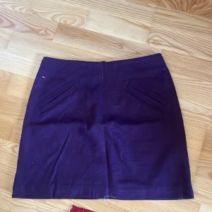 Mörklila kjol - Snygg mörklila kortkjol med en stilren design. Kjolen har två dekorativa fickor framtill och en figurnära passform. Perfekt för att ge en färgklick till din outfit!