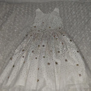 Vit klänning med stjärnor från H&M - Säljer en söt vit klänning från H&M med glittriga stjärnor i guld och silver. Klänningen är ärmlös och har en vid kjol som ger en fin volym. Perfekt för festliga tillfällen! 🌟