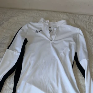 Vit långärmad tröja från Nike - Säljer en vit långärmad tröja från Nike med dragkedja vid halsen. Tröjan har svarta detaljer längs ärmarna och en liten Nike-logga på bröstet. Perfekt för sportiga aktiviteter eller en avslappnad stil.