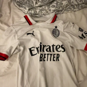 Vit AC Milan fotbollströja från Puma - Säljer en vit AC Milan fotbollströja från Puma med korta ärmar och röda detaljer. Tröjan har klubbens emblem på bröstet och sponsorlogga framtill. Perfekt för alla fans av laget!