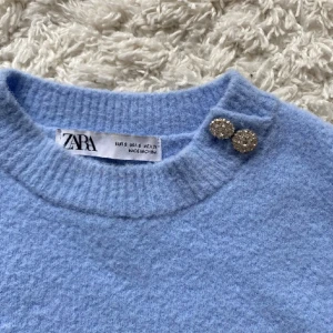 Ljusblå stickad tröja från Zara - Säljer en superfin ljusblå stickad tröja från Zara. Den har en rund halsringning och två glittriga knappar på axeln som en snygg detalj. Tröjan är kortärmad. (Lite  nopprig)