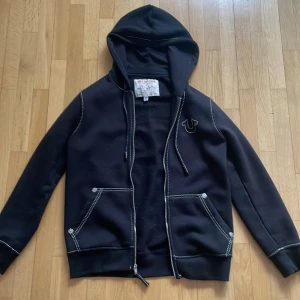 Svart  zip up hoodie från True Religion - Snygg svart zip up hoodie från True Religion med vit kontrastsöm och dragkedja. Den har en stor logga på ryggen och två fickor fram. Knappt andvänd, finns inga defekter. Den är lite  är overzied. Skriv om du har frågor eller mer bilder. Köpt för 1600kr