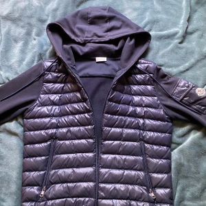 Moncler cardigan - Snygg svart cardigan från Moncler med huva och dragkedja perfekt till våren. Har en liten defekt på vänster sida men det syns knappt, hålet är även mindre än lagningen om man vill ta bort de. Storlek S och är använd en höst. Kan även tänka mig byta mot en annan cardigan,pris kan diskuteras vid snabb affär!