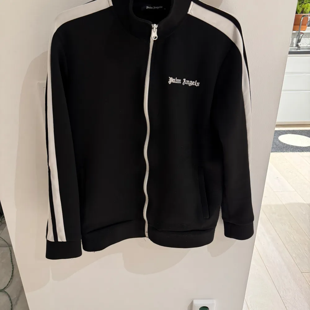 Snygg svart träningsjacka från Palm Angels med vita ränder längs ärmarna och dragkedja framtill. Jackan har en stilren design med broderad logga på bröstet. Perfekt för en sportig look! Koftan har en fungerande QR-kod också . Neuletakit & Villapaidat.