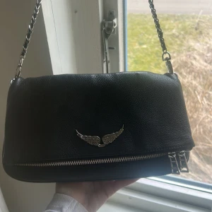 Ekta Zadig & volentaire väska  - Snygg svart axelväska i skinn med kedjedetaljer och en cool vingdekor på framsidan. Väskan har en dragkedja både fram och bak för extra säkerhet och stil. Perfekt för att ge din outfit en edgy touch. Säljer pga bytt stil så vill rensa