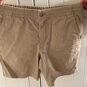 Beige shorts från Polo Ralph Lauren - Snygga beige shorts från Polo Ralph Lauren med elastisk midja och knappstängning. De har två bakfickor med knappar och en klassisk design som passar perfekt för varma dagar.