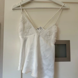 Vit satinlinne med spetsdetaljer från ZARA - Säljer ett elegant vitt satinlinne med tunna axelband och vackra spetsdetaljer vid bysten. Perfekt för en stilren look. Linnet har en mjuk och glansig yta som ger en lyxig känsla.