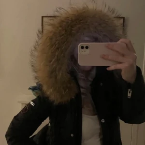 Svart parkajacka med pälsdetalj - Snygg svart parkajacka med stor pälsdetalj på huvan. Jackan har dragkedja och knappar framtill samt långa ärmar med dragkedjor vid ärmsluten. Perfekt för kyliga dagar.