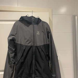 Svart och grå vindjacka från Haglöfs - Säljer en stilren svart och grå vindjacka från Haglöfs. Jackan har en praktisk huva och dragkedja framtill. Perfekt vårjacka men också  för blåsiga dagar och utomhusaktiviteter.